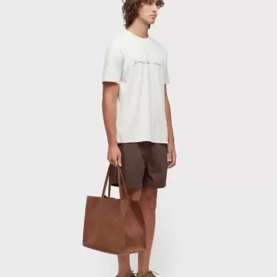 Homem com t-shirt branca, calções castanhos, sapatos castanhos e mala castanha em camurça