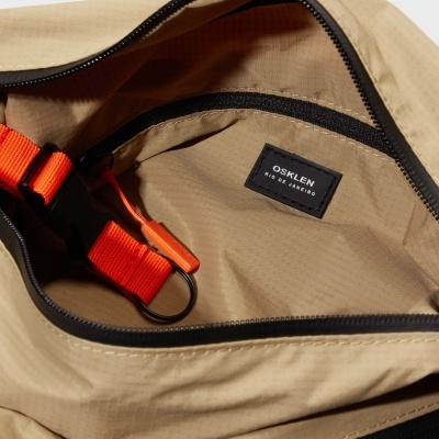 Mala bege clara com cinto vermelho e etiqueta OSKLEN