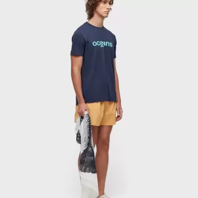Homem com t-shirt azul escura com texto 'oceans', calções amarelos e toalha branca preta.