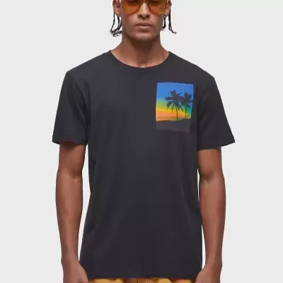 Homem com t-shirt preta com estampa de pôr do sol e palmeiras, calções brancos com manchas amarelas e óculos de sol castanhos