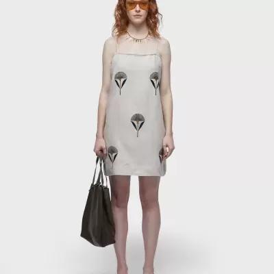 Modelo com vestido curto cinza claro estampado, óculos laranja, bolsa preta e sapatos bege em fundo branco