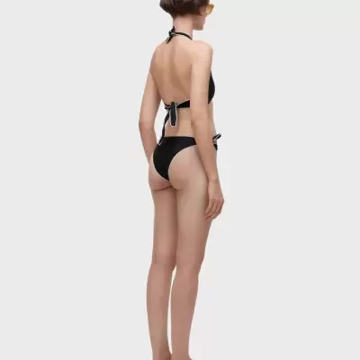 Mulher de costas a vestir biquíni preto com amarração e sandálias pretas com biqueira bege.