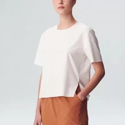 Mulher a usar t-shirt branca de manga curta e calças castanhas num fundo cinzento claro