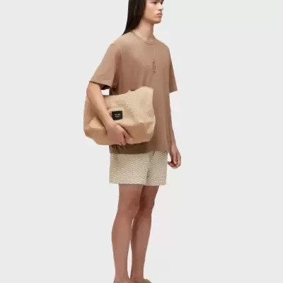 Pessoa com roupa castanha e bege segurando bolsa de palha com etiqueta negra