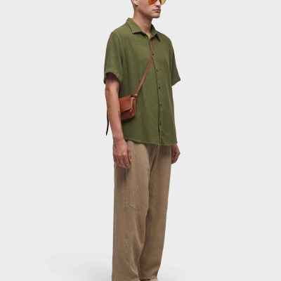 Homem vestido com camisa verde, calças castanhas largas e sapatos bege, com bolsa castanha pequena ao ombro.