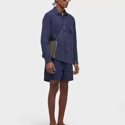 Homem vestido com camisa e calções azul-escuros, sandálias verdes, óculos escuros e bolsa cinza ao ombro