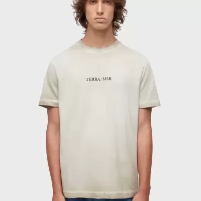 Homem a usar t-shirt bege com texto TERRA/MAR preta e calças azuis.