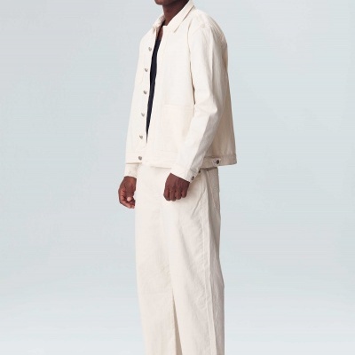 Homem com casaco e calças de ganga brancas e ténis brancos em fundo cinzento claro.