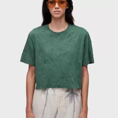 Mulher com t-shirt verde enrugada, calções bege com padrão e óculos de sol castanhos