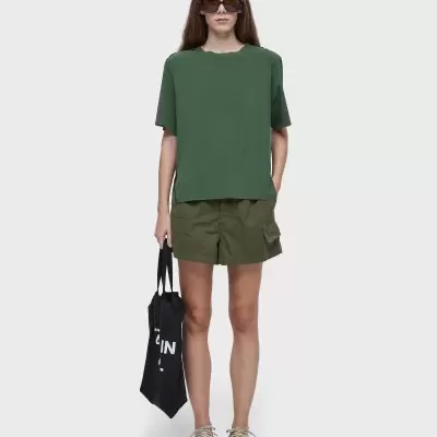 Mulher com t-shirt verde escuro, calções verdes, ténis bege, óculos escuros e tote bag preta com texto branco