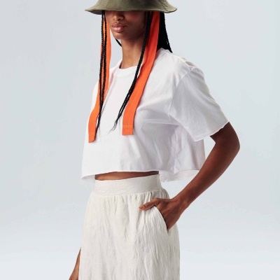 Mulher com chapéu verde, t-shirt branca curta e calças brancas largas.