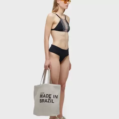 Mulher com fato de banho preto, óculos de sol amarelos e saco branco com texto 'MADE IN BRAZIL'