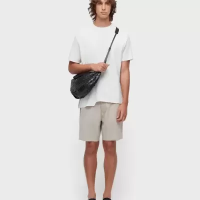 Homem com t-shirt branca, calções cinza claro, sapatos pretos e mala preta trançada numa parede branca.