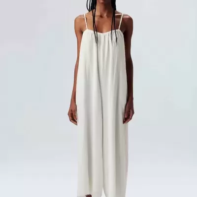 Mulher vestindo macacão branco longo com alças finas, chapéu bege e sandálias pretas em fundo cinza-claro