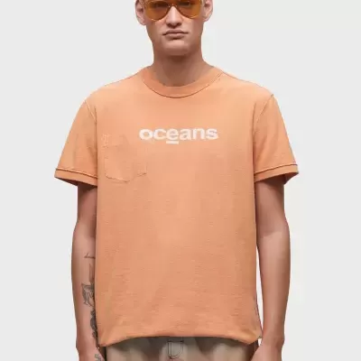 T-shirt laranja clara com texto 'Oceans' e bolso, usada por modelo com óculos.