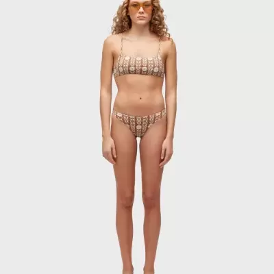 Mulher com biquíni bege com padrão floral branco, chapéu e óculos amarelos, e sandálias brancas em fundo cinzento.