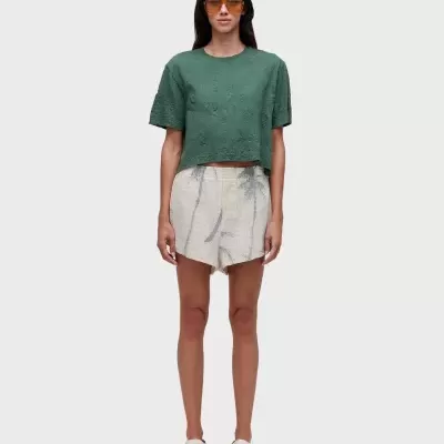 Mulher vestindo t-shirt verde, calções com padrão de folhas cinzentas e sandálias brancas com óculos de sol.