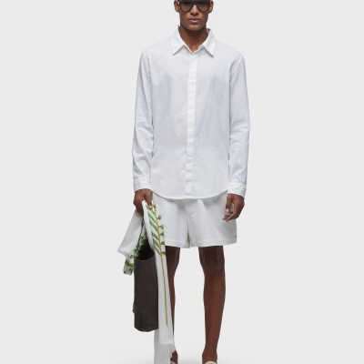 Homem com camisa branca, calções brancos, sapatilhas bege e óculos escuros segurando peças de roupa e bolsa preta