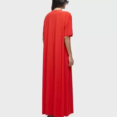 Vestido longo vermelho de manga curta na parte traseira, usado por modelo com óculos escuros e sapatilhas em fundo cinzento.