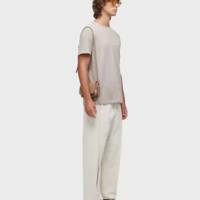 Homem com roupa casual clara e acessórios castanhos em fundo cinza.