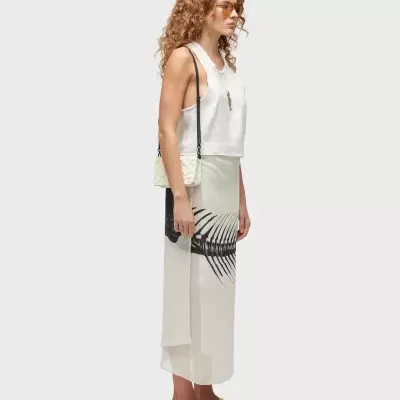 Mulher vestindo blusa branca, saia longa com padrão preto, sandálias brancas, mala bege e óculos de sol laranja.