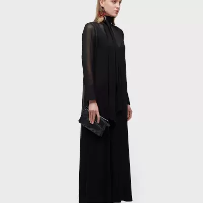 Mulher vestindo macacão preto de manga comprida transparente com gola alta e laço, segurando carteira preta.