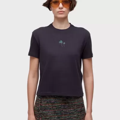 Mulher com t-shirt preta com bordado de palmeiras, saia curta colorida e óculos de sol âmbar.