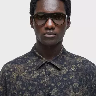 Homem de pele escura com camisa floral translúcida preta e óculos castanhos largos.
