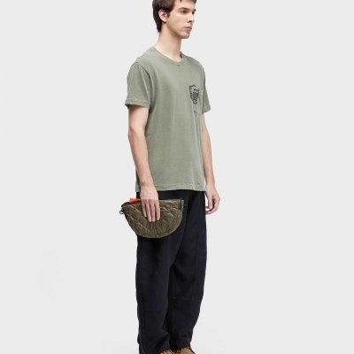 Homem vestido com t-shirt verde caqui, calças negras, sapatos castanhos, segurando bolsa verde almofadada