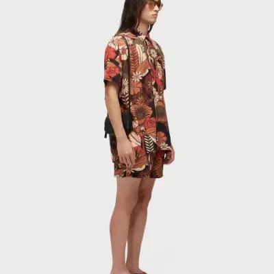 Homem com conjunto estampado floral laranja e castanho, sandálias castanhas, óculos âmbar e bolsa preta