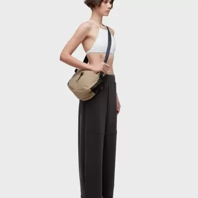 Mulher com top branco, calças pretas largas, ténis brancos e mochila bege com detalhes pretos da marca OSKLEN.