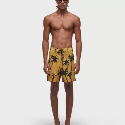 Modelo masculino com calções de banho castanhos com padrão de palmeiras pretas, óculos escuros e chinelos pretos