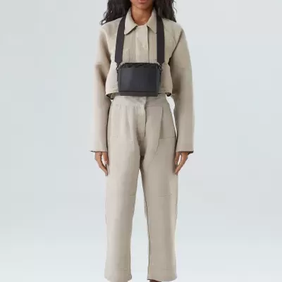 Mulher com macacão bege e mala preta pequena ao peito em fundo cinza claro