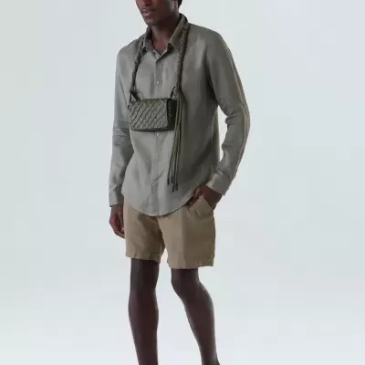 Homem com camisa cinza, calções bege, sapatilhas castanhas e bolsa verde ao pescoço