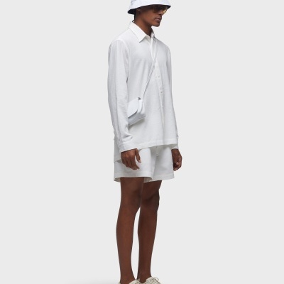 Homem vestido com camisa branca, calções, chapéu e tênis brancos, bolsa pequena branca pendurada no ombro.