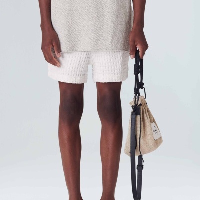 Pessoa com bermuda branca texturizada, sandálias pretas e bolsa bege de tecido com alça preta