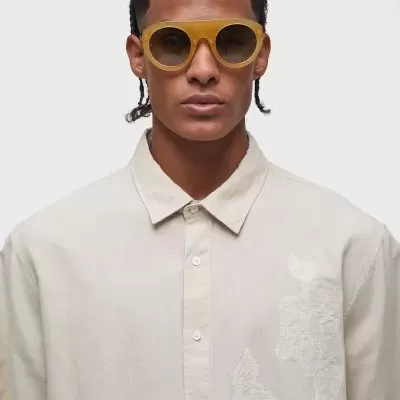 Homem com óculos de sol amarelos e camisa bege clara com bordado branco