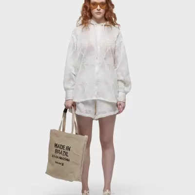 Mulher com conjunto branco de hoodie e calções, óculos laranja, e sacola de tecido bege com texto preto.