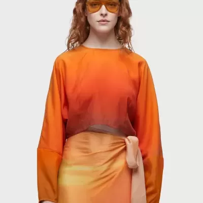 Mulher com blusa laranja degradê, saia laranja e amarelo com laço, óculos de sol cor âmbar