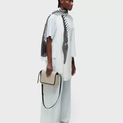 Modelo com roupa branca, lenço preto e branco, e mala bege com detalhes pretos.