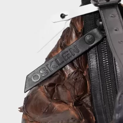 Detalhe de mochila de pele castanha com zipper e tira preta com texto OSKLEN