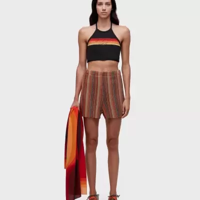 Mulher com top cropped com riscas coloridas e calções riscas verticais multicoloridas, ténis laranja e castanho, segurando peça de tecido colorido