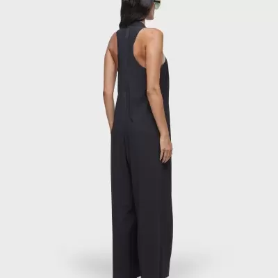 Mulher de costas veste macacão preto e sandálias pretas com óculos de sol