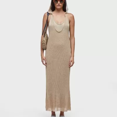 Mulher com vestido longo bege de malha e bolsa trançada, usando óculos de sol e sandálias brancas