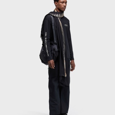 Homem com roupa casual preta, echarpe padrão bege, bolsa e ténis pretos num fundo branco.