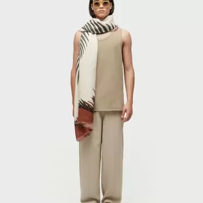 Modelo veste regata e calças bege claro, lenço branco com padrão de folhas, mocassins castanhos e óculos de sol amarelos