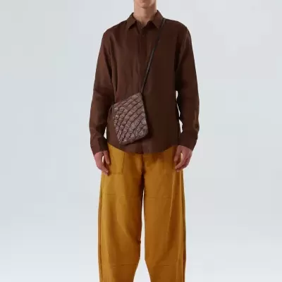 Homem com camisa castanha, calças mostarda largas, sapatos castanhos e bolsa pequena matelassê