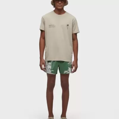 Homem com camisa bege, calções verdes com padrão branco, chapéu branco e ténis bege