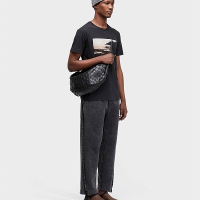 Homem de lado com t-shirt preta, calças cinza, gorro cinza e mala preta com padrão em escamas, sapatos castanhos e pretos