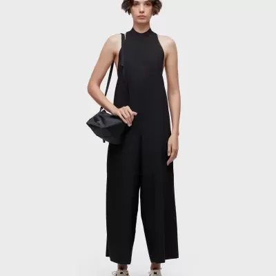 Mulher com macacão preto largo, ténis brancos e bolsa preta pequena em fundo cinzento claro.